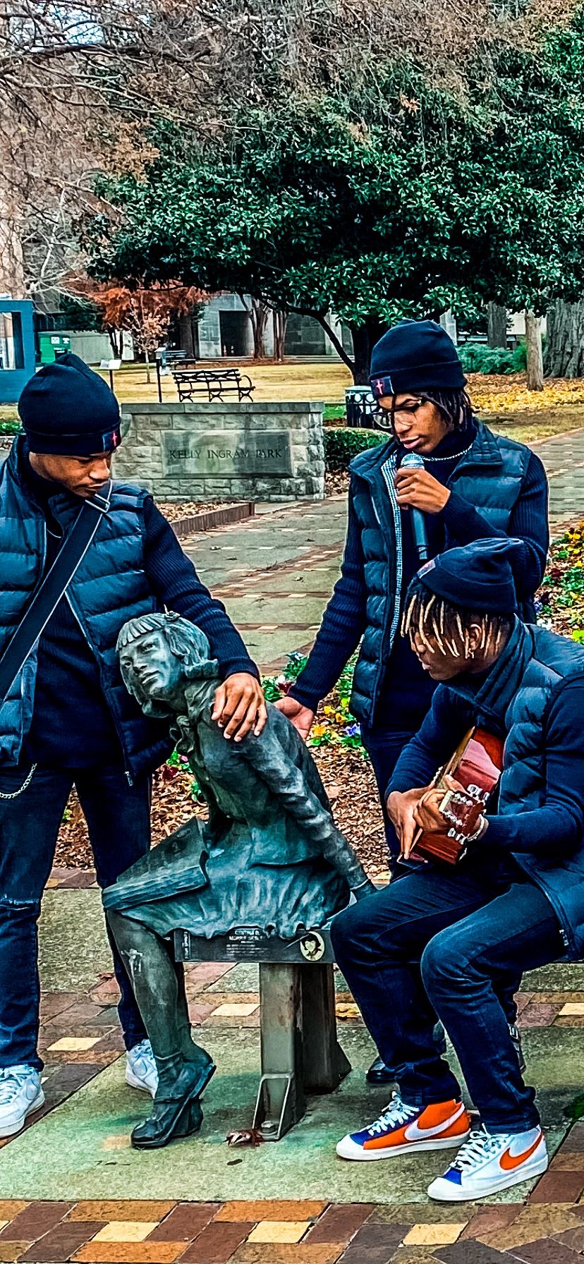 Three young men interact with a statue while one plays guitar in a park.