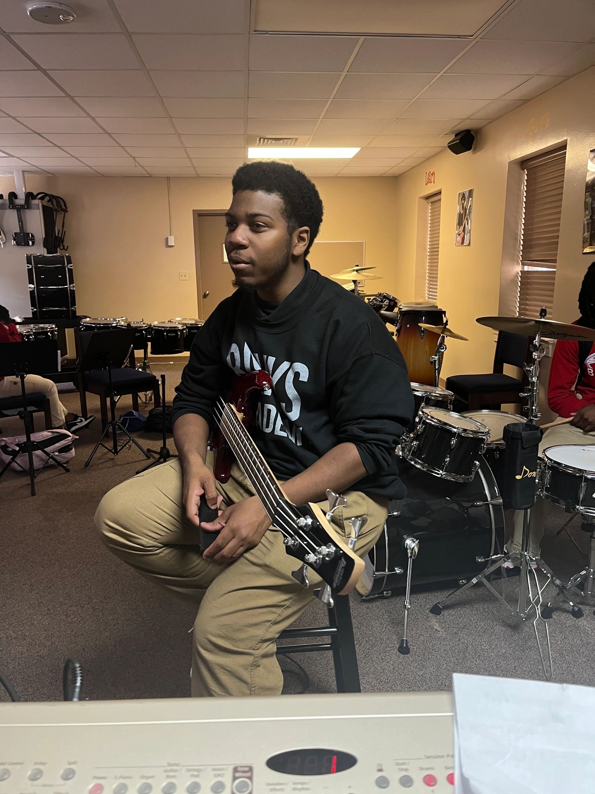 Young man sitting on a stool holding a bass guitar in a music room.