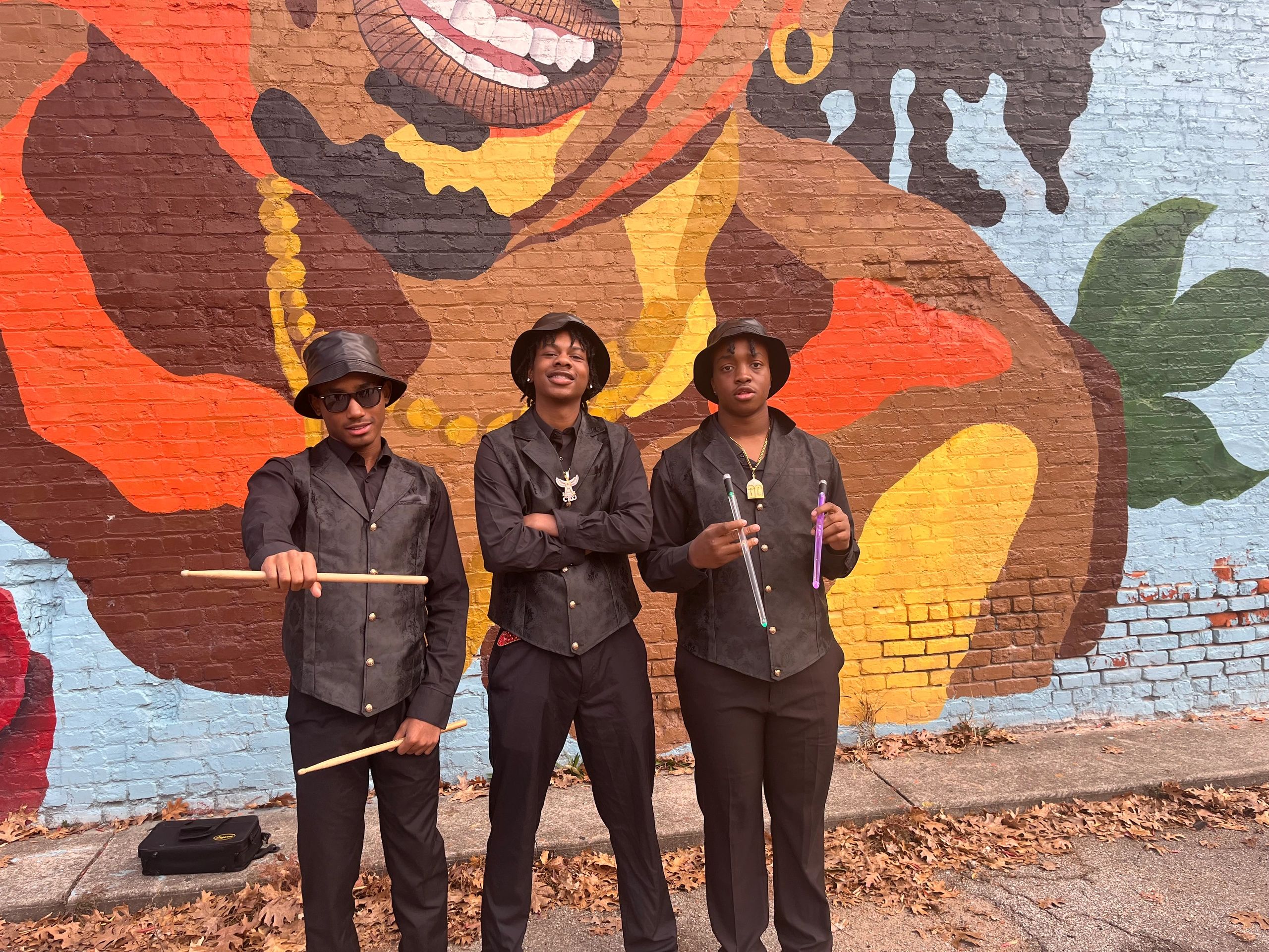 Three young men in black attire posing with drumsticks in front of vibrant mural.