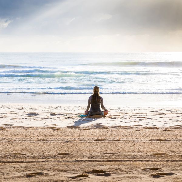 Person meditating alone on a sandy beach facing the ocean waves.