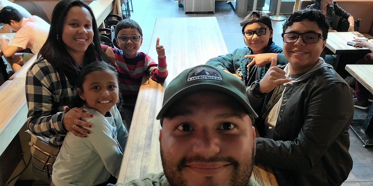 A happy group selfie of six people gathered around a wooden table in a casual dining setting.