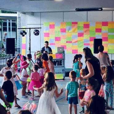 Children dancing at a colorful party with a DJ in the background.