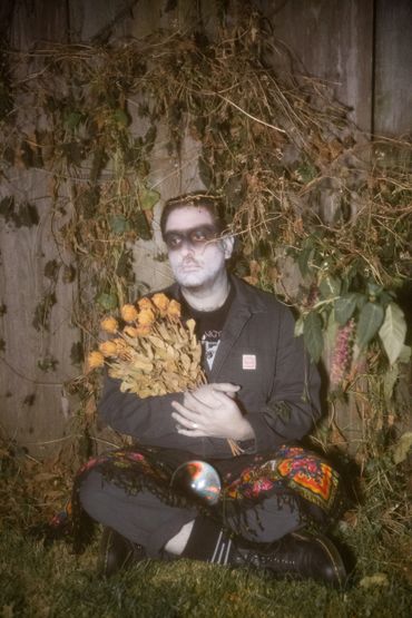a dream ghoul sits in front of a viney fence, holding a bouquet of dead roses and a crystal ball.