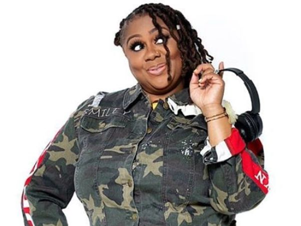 Woman in camo jacket with headphones over her shoulder, smiling playfully.