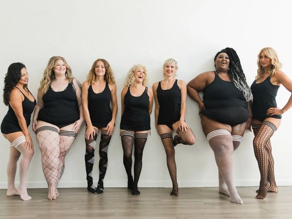 Diverse women in black outfits and patterned stockings posing against a white wall.