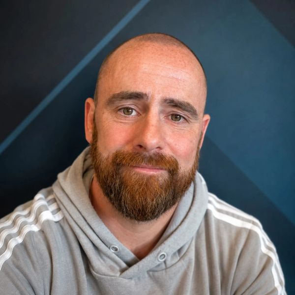 Smiling bald man with a beard wearing a grey hoodie against a dark geometric background.