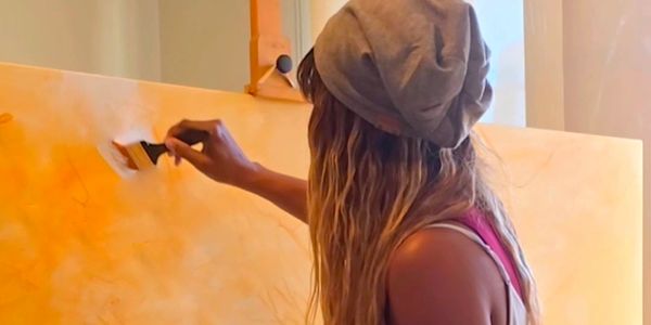Woman painting a canvas with a brush, wearing a gray beanie and pink top.
