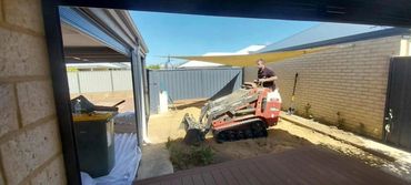 Man operating a small tracked excavator in a backyard under clear blue sky.