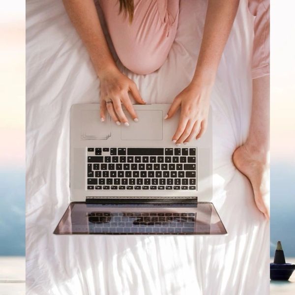 Person using a laptop while sitting on a bed in pink pajamas.