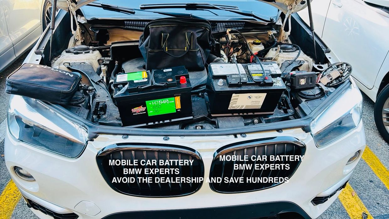 Open hood of white BMW showing two car batteries and promotional text on grille.