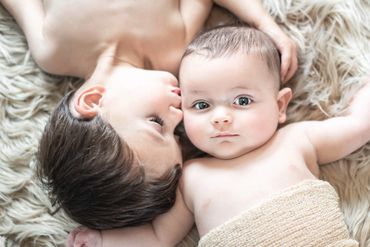 Young boy kissing his newborn baby brother on the cheek posed on a neutral fluffy rug