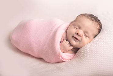 Newborn baby girl wrapped up in a soft pink blanket, sleeping an smiling, hand under her chin