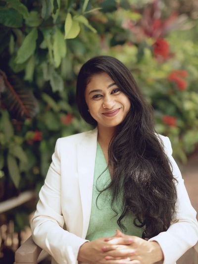 A woman smiling into the camera. She is wearing a green blouse and white jacket. 