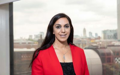 Woman in a red blazer smiling indoors with cityscape background.