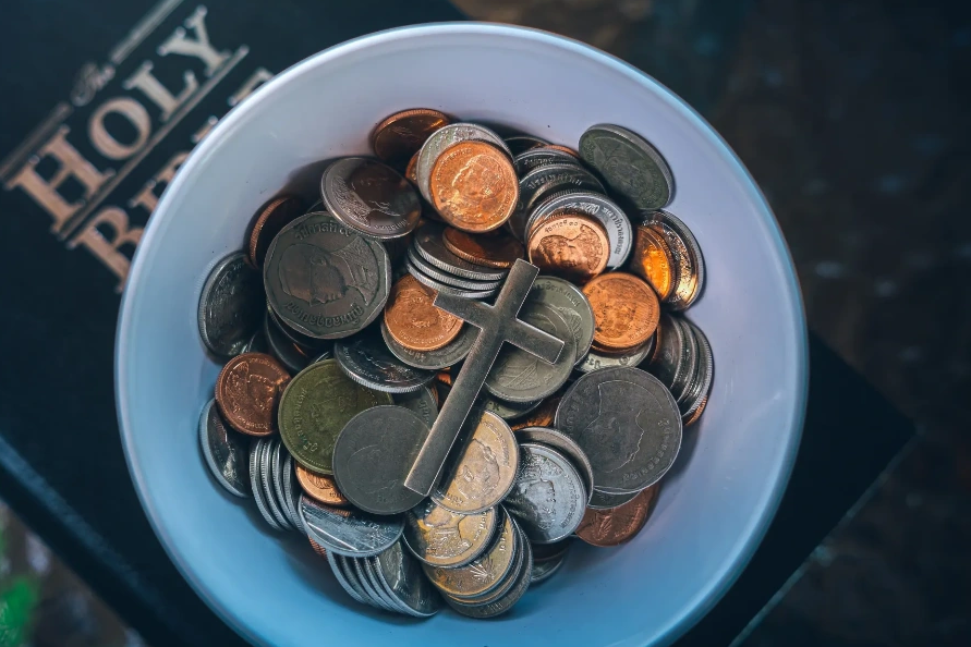 A cross on top of coins inside a white bowl with a Holy Bible in the background.