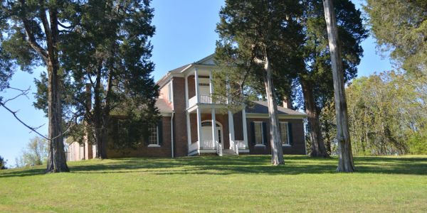 Historic brick house surrounded by tall trees and green lawn.
