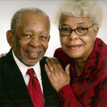 Elderly couple smiling warmly, dressed formally against a plain background.