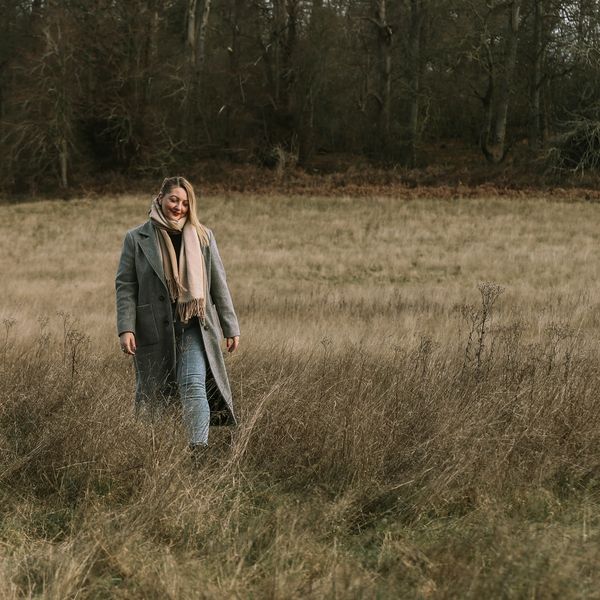A woman with blonde hair walking towards the camera in the woods in a grey coat and beige scarf
