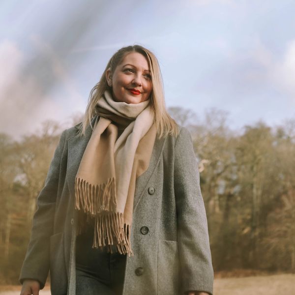 A woman with blonde hair and red lipstick in a beige scarf and grey coat standing in the woods smili