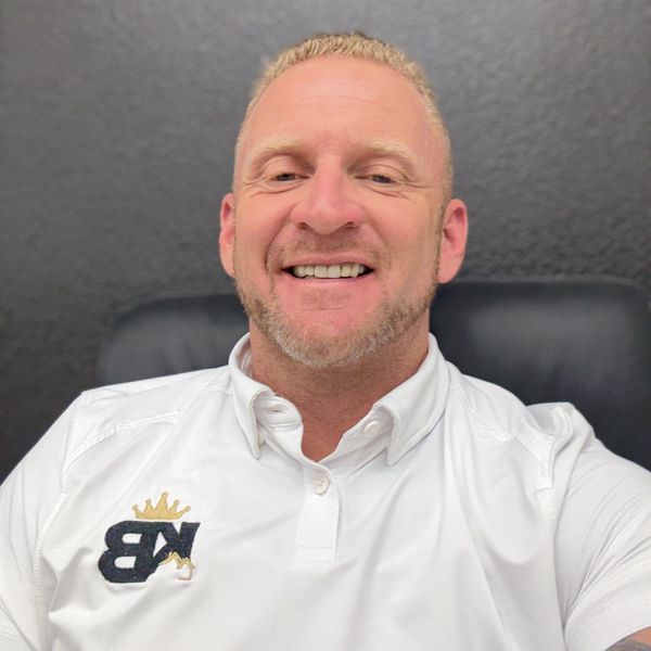 Smiling man in a white shirt with a logo, sitting against a gray wall.