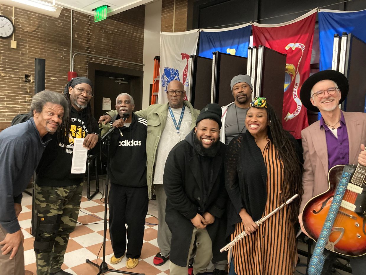 A diverse group posing indoors, one holding a guitar, another a flute, with flags in the background.