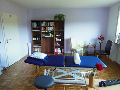 Praxisraum mit blauer Behandlungsliege, Bücherregal und netter Sitzgruppe mit Blumen