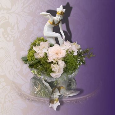 Delicate porcelain dancer figurine atop floral arrangement in reflective glass bowl.