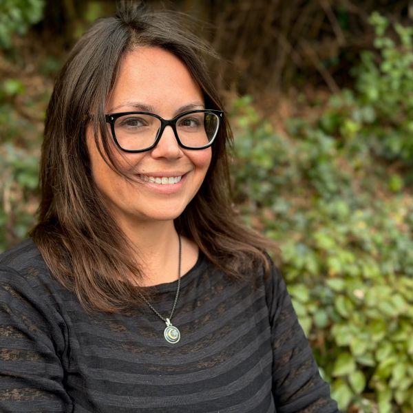 Smiling woman with glasses outdoors, wearing a striped top and pendant necklace.