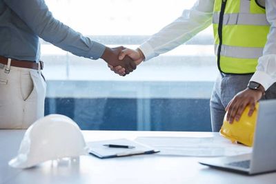 Two professionals shaking hands over a desk with safety helmets and documents.