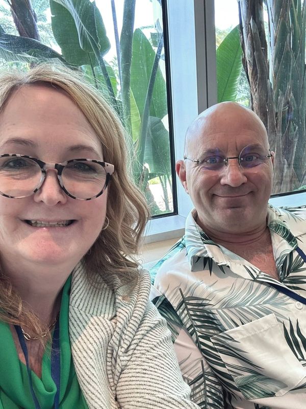 Smiling man and woman sitting indoors with tropical plants outside the window.