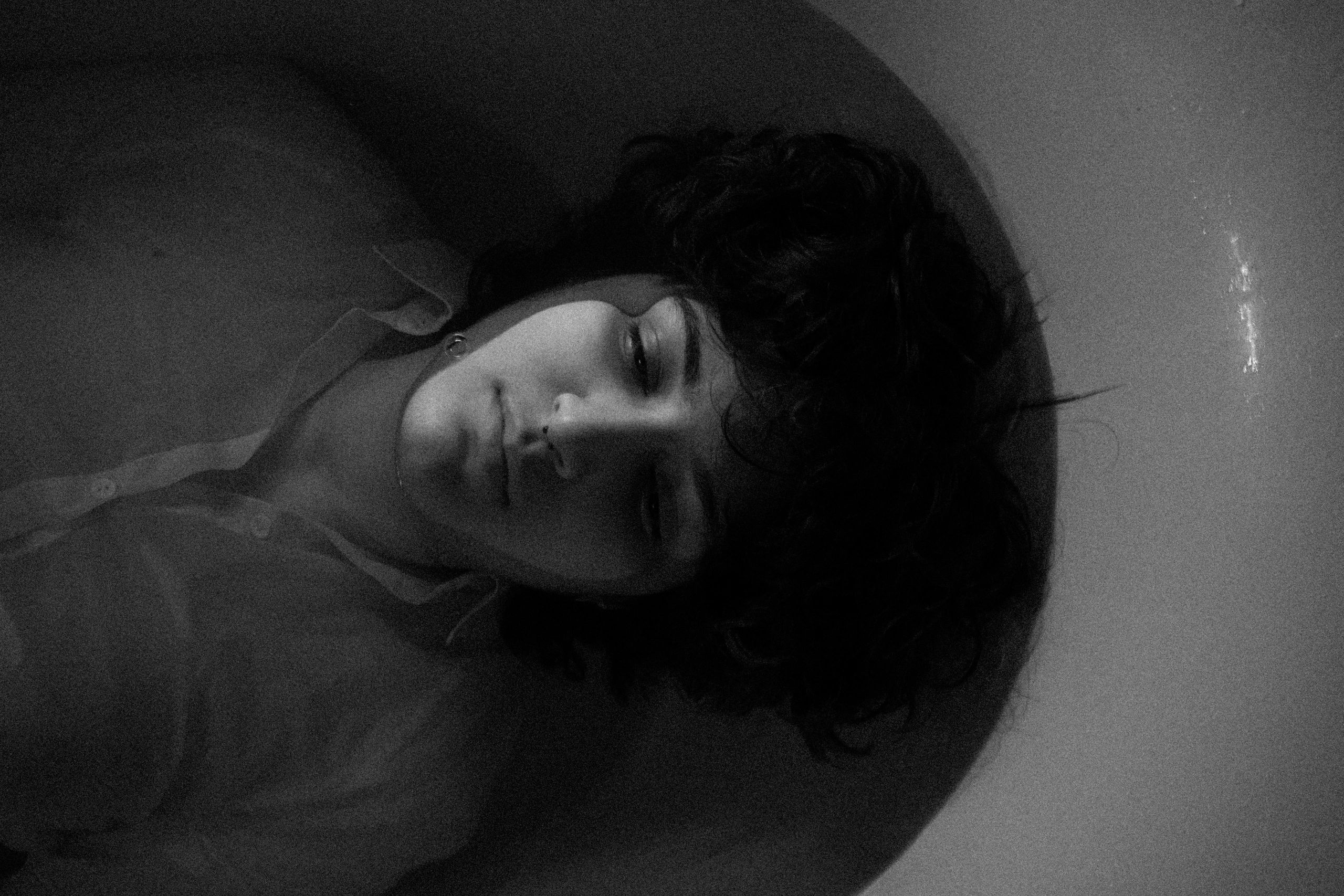 Black and white portrait of a person lying in water, eyes closed, with curly hair and a nose piercing.