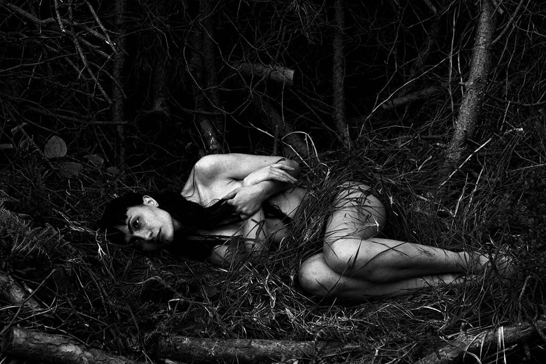 A woman lies curled up naked in a dark, dense forest surrounded by branches and grass.