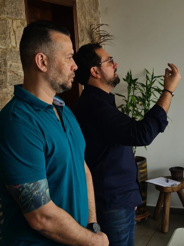 Two men standing indoors, one with tattoos and the other gesturing.