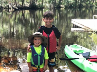 Two boys in life jackets standing by a kayak on a calm lake.