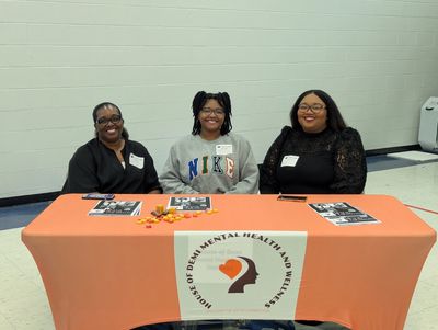 Three women sit behind a mental health awareness table with informational flyers and candies.