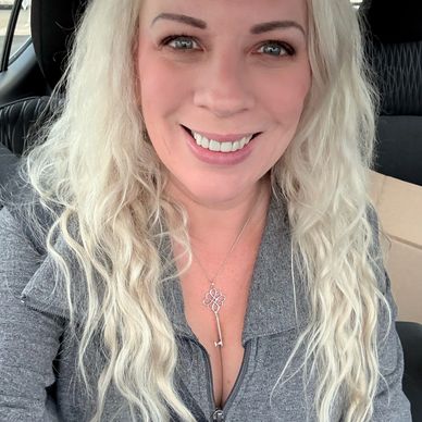 Smiling woman with long blonde hair wearing a gray zip-up sweater and a key necklace.