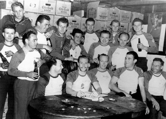 Group of men in matching shirts with beer bottles, posing around a table.