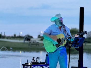 Man in cowboy hat plays guitar outdoors near water.