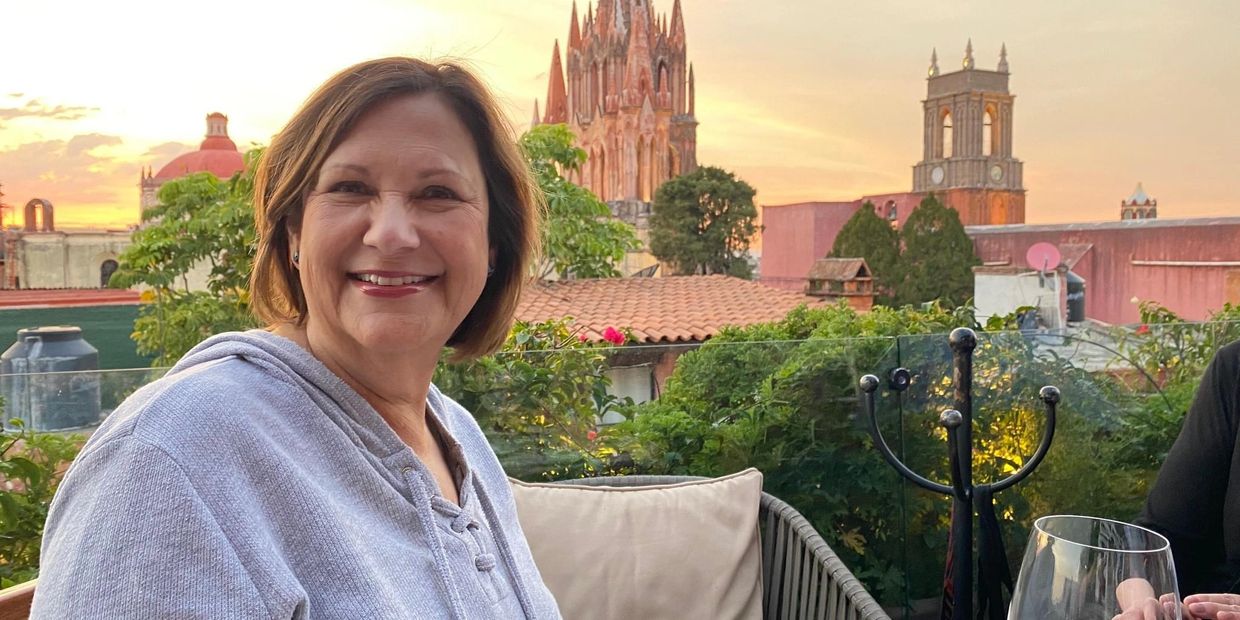 Smiling woman in a gray hoodie at a scenic rooftop restaurant during sunset.