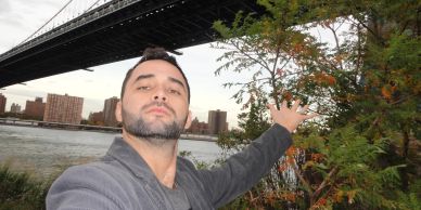 Man taking a selfie under a large bridge near water and trees.