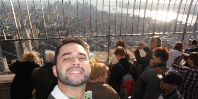 Man takes a selfie with city skyline and tourists behind a safety fence.