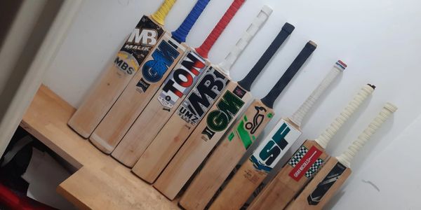 A row of cricket bats lined up on a wooden bench against a white wall.