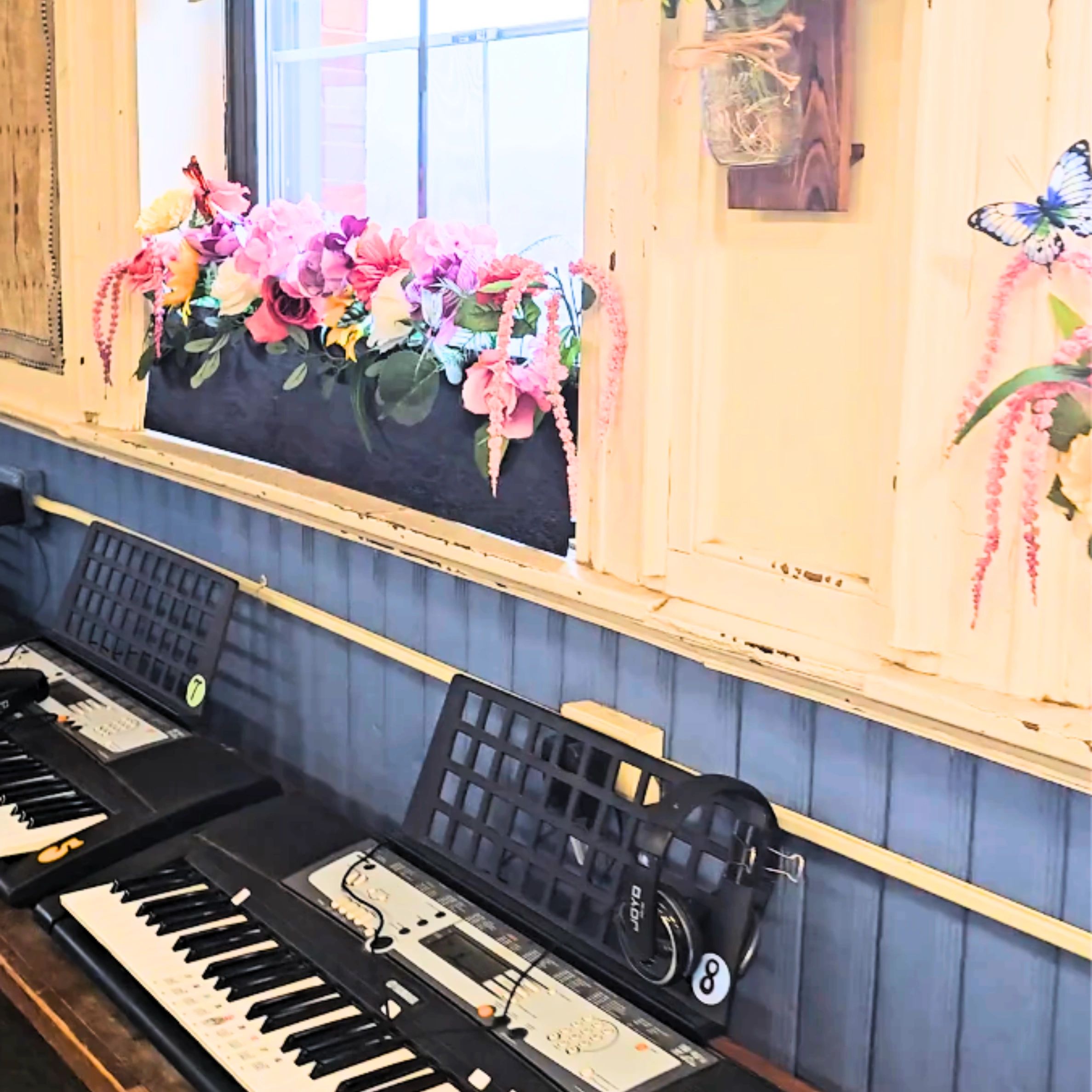Two keyboards with headphones against a blue wall under a flower-adorned window.