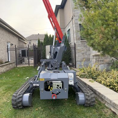 A compact tracked robot with a red arm attachment on grass beside a stone house.