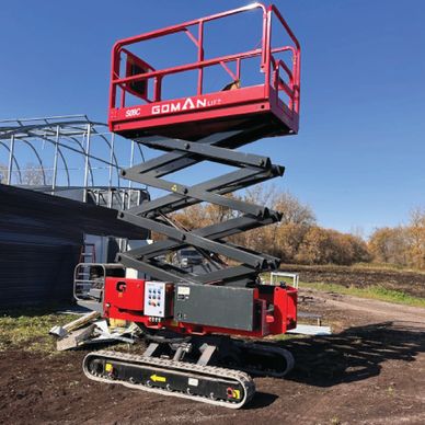 Red scissor lift with tracked base outdoors on dirt ground under clear blue sky.
