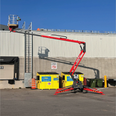 A red cherry picker lift extended beside a building with yellow dumpsters.