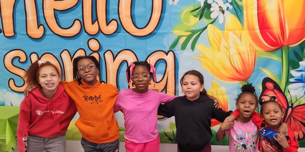 Six children smiling and posing in front of a colorful spring banner.