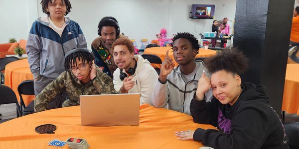 Group of six young people gathered around a laptop at an orange-covered table.