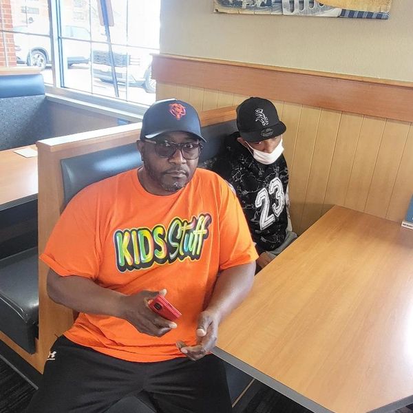 Two men sitting in a restaurant booth, one wearing an orange "KIDS Stuff" shirt.