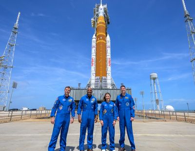 Four astronauts in blue suits stand before a large rocket on a clear day.